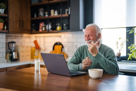 Téléchargez les photos : Homme âgé connecté sur Internet avec un ordinateur portable à la maison. Homme âgé avec une tasse de thé en utilisant un ordinateur portable à la table dans la cuisine. Homme mûr tapant sur ordinateur portable dans la cuisine pendant le petit déjeuner et le café driking. - en image libre de droit