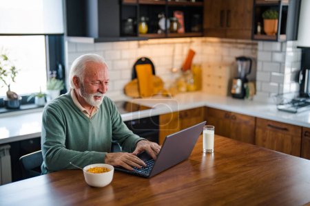 Photo for Busy with work. Grey-haired bearded senior man eyeglasses looking concentrated. One senior businessman standing in the kitchen at home and working on the laptop while drinking coffee. - Royalty Free Image