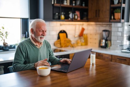 Téléchargez les photos : Homme avec ordinateur à la maison dans la cuisine moderne. Homme âgé connecté sur Internet avec un ordinateur portable à la maison. Télétravail à la retraite. Homme âgé souriant utilisant un ordinateur portable dans la cuisine à la maison - en image libre de droit