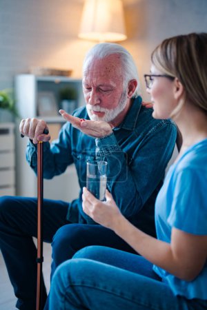 Photo for Caregiver or nurse giving and showing a pill to senior man on the table. Doctor or nurse caregiver showing a prescrption drug to senior man at home or nursing home. Giving the medicine - Royalty Free Image