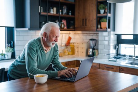 Photo for Senior man working at laptop at home. Retired Man On Phone At Home In Kitchen Using Laptop Celebrating Good News. Senior adult in home kitchen with tea cup and laptop. - Royalty Free Image