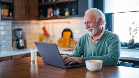 Téléchargez les photos : Homme âgé joyeux pendant la vidéoconférence dans la cuisine sur l'ordinateur portable tout en appréciant le petit déjeuner et une tasse de café. Personne âgée utilisant Internet chat en ligne technologie webcam vidéo faire un appel vidéo - en image libre de droit
