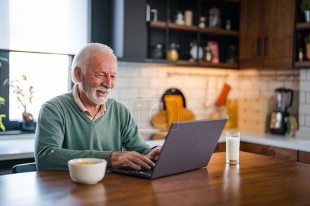 Foto de Hombre mayor conectado a Internet con el ordenador portátil en casa. Hombre mayor con taza de té usando portátil en la mesa en la cocina. Hombre maduro escribiendo en el ordenador portátil en la cocina durante el desayuno y el café goteo. - Imagen libre de derechos