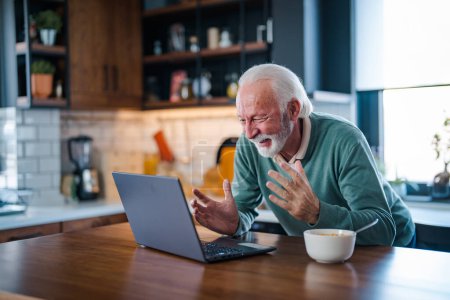Téléchargez les photos : Homme âgé joyeux pendant la vidéoconférence dans la cuisine sur l'ordinateur portable tout en appréciant le petit déjeuner et une tasse de café. Personne âgée utilisant Internet chat en ligne technologie webcam vidéo faire un appel vidéo - en image libre de droit
