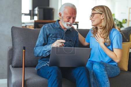 Photo for Friendly pretty young woman nurse assisting disabled senior man at home using laptop, sitting at desk in front of computer, having conversation and smiling, man touching gadget screen - Royalty Free Image