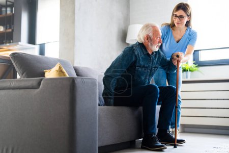 Photo for Smiling nurse assisting senior man to get up from bed. Caring nurse supporting patient while getting up from bed and move. Helping elderly disabled man standing up in his living room. - Royalty Free Image