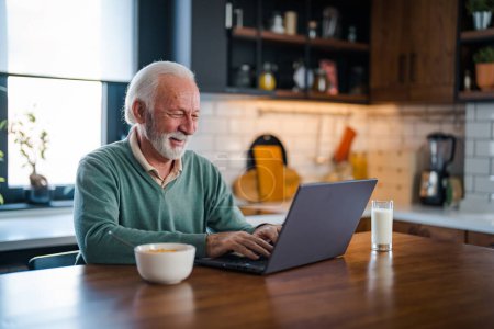 Photo for One senior businessman standing in the kitchen and working on the laptop  while drinking coffee. Grey-haired bearded senior man eyeglasses looking concentrated - Royalty Free Image