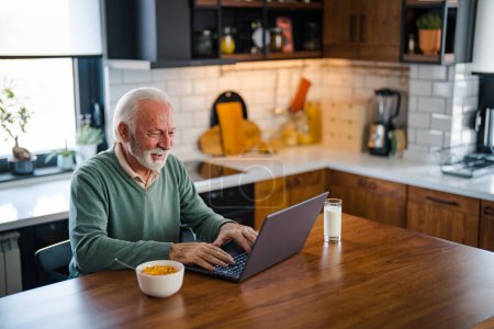 Foto de Hombre con ordenador en casa en la cocina moderna. Hombre mayor conectado a Internet con el ordenador portátil en casa. Trabajo remoto en la jubilación. Hombre mayor sonriente usando el ordenador portátil en la cocina en casa - Imagen libre de derechos