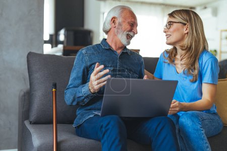 Photo for Senior man with disability learning to pay bills online with laptop together with social worker. Nurse help senior retirement man working with laptop. Active senior man using laptop with female carer sitting by on sofa at home - Royalty Free Image