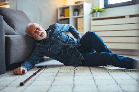 Photo for Elderly Senior Man Slip And Fall. Fallen Old Person in the Living Room. Sad tired stylish old man wearing checked shirt making effort trying to get up leaning on cane and divan in white light modern interior - Royalty Free Image