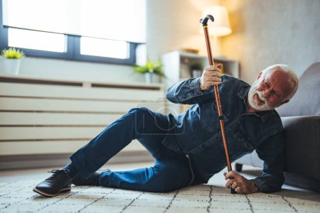 Téléchargez les photos : Un homme âgé qui utilise un bâton de marche est allongé sur le sol en bois dans son appartement après qu'il est tombé dans un accident grave. Homme âgé couché sur le sol de sa chambre dans une maison de retraite assistée - en image libre de droit