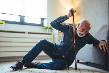 Téléchargez les photos : Blessé homme âgé gémissant de douleur alors qu'il essaie de se lever avec un bâton après la chute à la maison. Il se tient le bas du dos et peut à peine se lever. Homme âgé tombé sur le tapis avec bâton de marche - en image libre de droit