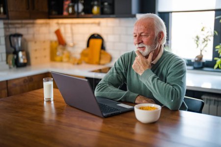 Téléchargez les photos : Télétravail à la retraite. Homme âgé souriant utilisant un ordinateur portable dans la cuisine à la maison, bel homme âgé dans les lunettes de vue regardant l'écran d'ordinateur et prenant des notes sur le bloc-notes, l'espace de copie - en image libre de droit