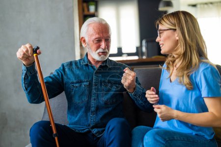 Téléchargez les photos : Jeune infirmière parlant avec un homme âgé avec un bâton handicapé assis sur le canapé à la maison - en image libre de droit