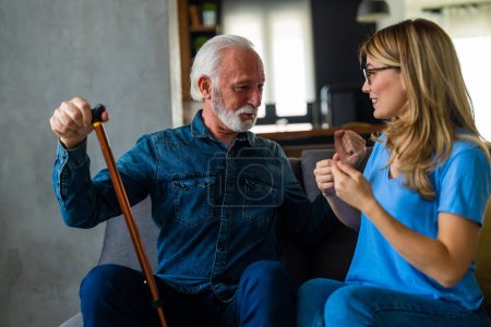 Photo for Young female nurse talking with senior man with disabled stick sitting on sofa at home - Royalty Free Image