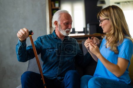 Photo for Young female nurse talking with senior man with disabled stick sitting on sofa at home - Royalty Free Image