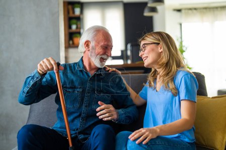Téléchargez les photos : Jeune infirmière et homme âgé avec un bâton handicapé assis et riant sur le canapé à la maison - en image libre de droit