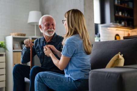 Téléchargez les photos : Jeune infirmière tenant la main d'un homme âgé avec un bâton handicapé sur le canapé à la maison - en image libre de droit