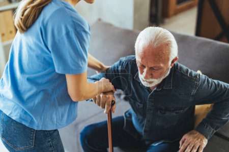 Téléchargez les photos : Infirmière aidant l'homme âgé handicapé à tenir debout du canapé à la maison - en image libre de droit