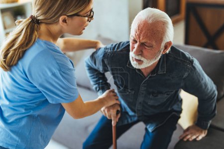 Photo for Nurse helping senior man with disabled stick standing up from sofa at home - Royalty Free Image