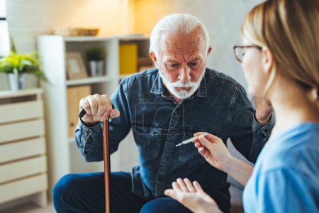 Téléchargez les photos : Jeune infirmière avec thermomètre parlant avec un homme âgé avec un bâton handicapé sur le canapé à la maison - en image libre de droit