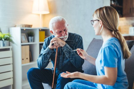 Foto de Enfermera joven con termómetro hablando con el hombre mayor con palo discapacitado en el sofá en casa - Imagen libre de derechos