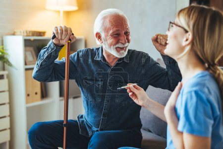 Téléchargez les photos : Jeune infirmière avec thermomètre parlant avec un homme âgé avec un bâton handicapé sur le canapé à la maison - en image libre de droit