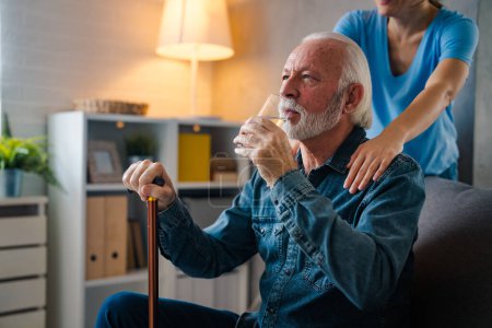 Téléchargez les photos : Infirmière partielle tenant la main sur les épaules d'un homme âgé avec un bâton handicapé buvant de l'eau du verre sur le canapé à la maison - en image libre de droit