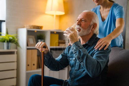 Foto de Enfermera parcial cogida de la mano sobre los hombros del hombre mayor con palo discapacitado beber agua de vidrio en el sofá en casa - Imagen libre de derechos