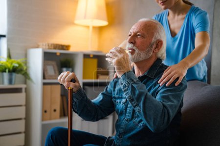 Photo for Partial nurse holding hands on shoulders of senior man with disabled stick drinking water from glass on sofa at home - Royalty Free Image