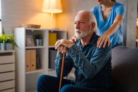 Téléchargez les photos : Infirmière cultivée tenant la main sur les épaules de l'homme âgé pensant avec un bâton handicapé sur le canapé à la maison - en image libre de droit