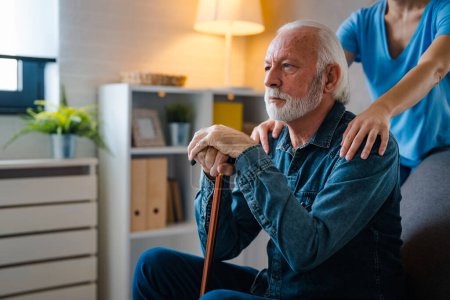 Foto de Enfermera recortada cogida de la mano sobre hombros de hombre mayor pensante con palo discapacitado en el sofá en casa - Imagen libre de derechos