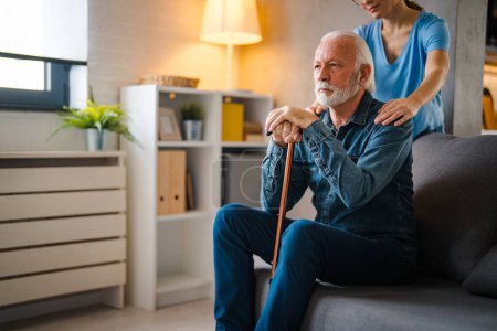 Cropped nurse holding hands on shoulders of thinking senior man with disabled stick on sofa at home