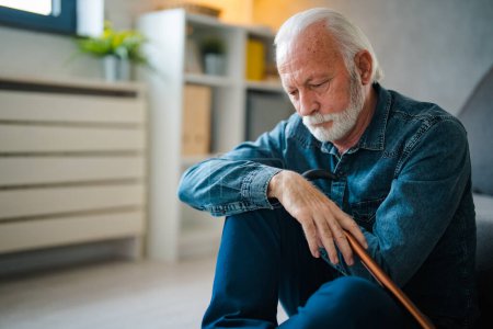 Photo for Sadness senior man with disabled stick sitting on floor near sofa at home - Royalty Free Image