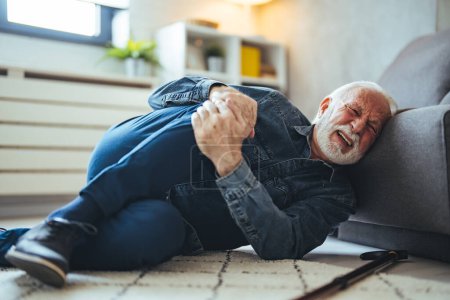 Foto de Hombre caucásico mayor acostado en el suelo y sufriendo de dolor cerca de sofá en casa - Imagen libre de derechos