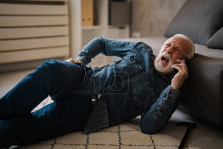 Foto de Maduro hombre caucásico con dolor tumbado en el suelo y hablando en el teléfono inteligente cerca de sofá en casa - Imagen libre de derechos