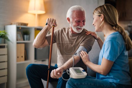 Téléchargez les photos : Jeune infirmière mesurant la pression d'un homme âgé avec un bâton handicapé sur le canapé à la maison - en image libre de droit