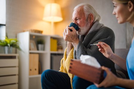 Foto de Enfermera joven con termómetro y servilletas y hombre mayor enfermo bebiendo té caliente en el sofá en casa - Imagen libre de derechos