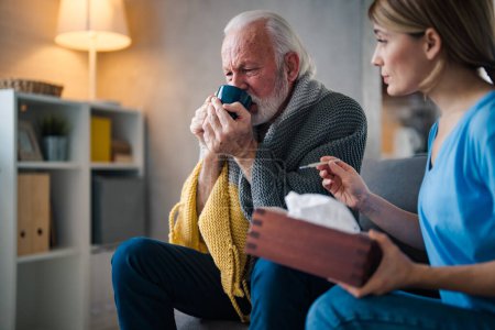 Foto de Enfermera joven con termómetro y servilletas y hombre mayor enfermo bebiendo té caliente en el sofá en casa - Imagen libre de derechos