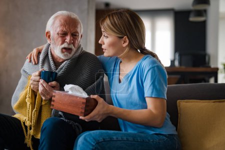 Foto de Enfermera joven con servilletas cuidando y abrazando la enfermedad hombre mayor bebiendo té caliente en el sofá en casa - Imagen libre de derechos
