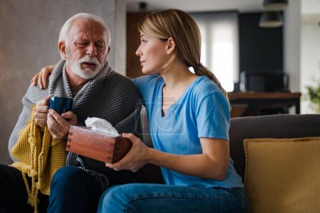 Foto de Enfermera joven con servilletas cuidando y abrazando la enfermedad hombre mayor bebiendo té caliente en el sofá en casa - Imagen libre de derechos