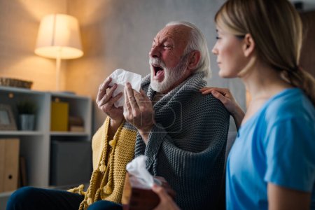Photo for Sickness mature man sneezing in napkin during sitting on sofa near young female nurse at home - Royalty Free Image
