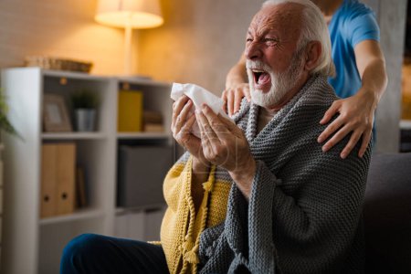 Photo for Partial nurse holding hands on shoulders of sickness senior man sneezing in napkin on sofa at home - Royalty Free Image