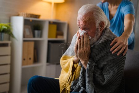 Photo for Partial nurse holding hands on shoulders of sickness senior man sneezing in napkin on sofa at home - Royalty Free Image