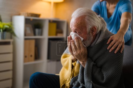 Téléchargez les photos : Infirmière partielle tenant la main sur les épaules d'un aîné malade éternuant dans une serviette sur un canapé à la maison - en image libre de droit