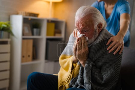 Foto de Enfermera parcial cogida de la mano sobre los hombros de la enfermedad hombre mayor estornudando en servilleta en el sofá en casa - Imagen libre de derechos