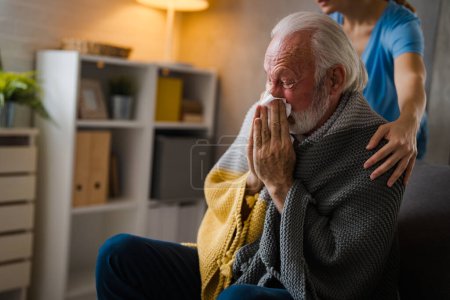 Foto de Enfermera parcial cogida de la mano sobre los hombros de la enfermedad hombre mayor estornudando en servilleta en el sofá en casa - Imagen libre de derechos