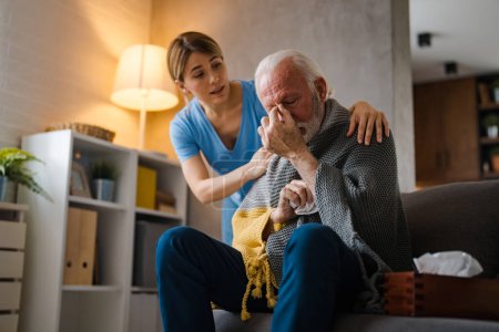 Téléchargez les photos : Jeune infirmière soignante de la maladie homme âgé avec serviette sur le canapé à la maison - en image libre de droit