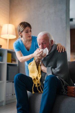 Téléchargez les photos : Jeune infirmière s'occupant de la maladie vieil homme éternuant dans la serviette sur le canapé à la maison - en image libre de droit