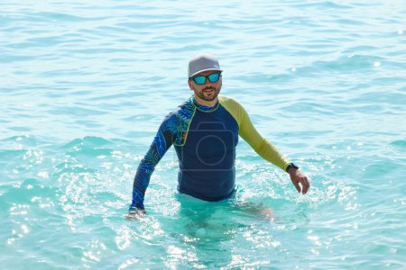 Photo for Handsome man with beard and mustache in a lycra shirt standing waist-deep in blue sea water. - Royalty Free Image